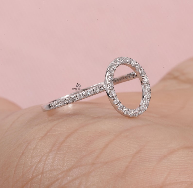 May include: A silver ring with a circular design, encrusted with small, sparkling stones. The band and the circle are both adorned with the same stones, creating a continuous, elegant look. The ring is set against a soft pink background.