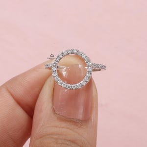 May include: A silver ring with a circular design, encrusted with small, sparkling stones. The ring features a halo of stones around the open circle, with additional stones along the band. The ring is held against a pink background.