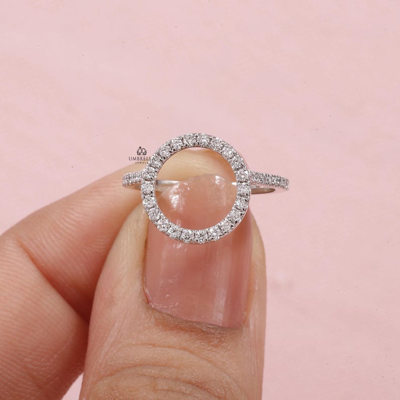 May include: A silver ring with a circular design, encrusted with small, sparkling stones. The ring features a halo of stones around the open circle, with additional stones along the band. The ring is held against a pink background.