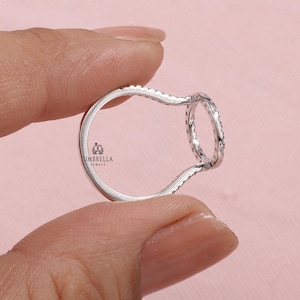 May include: A silver ring with a unique open design, held between fingers against a pink background. The ring features a continuous band of small, sparkling stones around a circular element. The brand "UMBRELLA JEWELS" is visible.