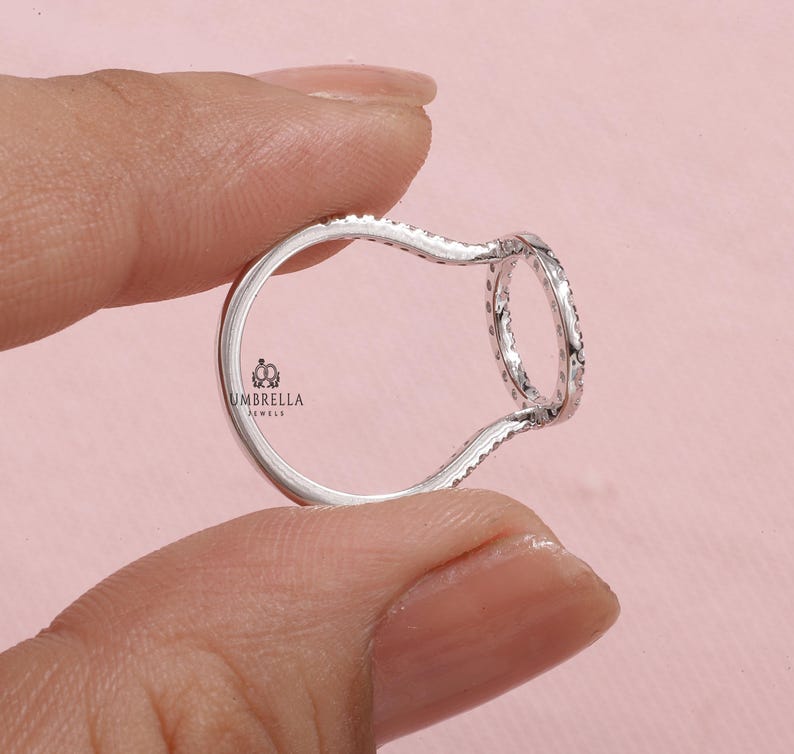 May include: A silver ring with a unique open design, held between fingers against a pink background. The ring features a continuous band of small, sparkling stones around a circular element. The brand "UMBRELLA JEWELS" is visible.
