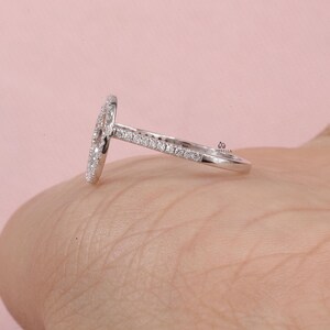 May include: A silver ring with a halo design, featuring a central oval-shaped cluster of small, clear stones. The band is also adorned with smaller stones, creating a sparkling effect. The ring is set against a soft pink background.