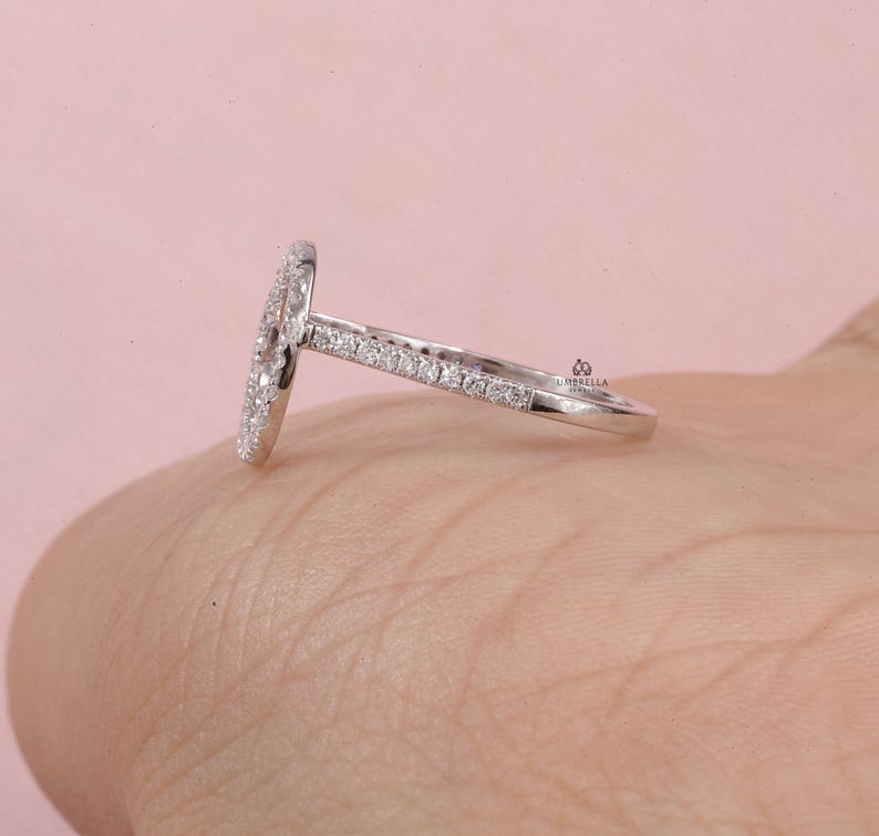 May include: A silver ring with a halo design, featuring a central oval-shaped cluster of small, clear stones. The band is also adorned with smaller stones, creating a sparkling effect. The ring is set against a soft pink background.