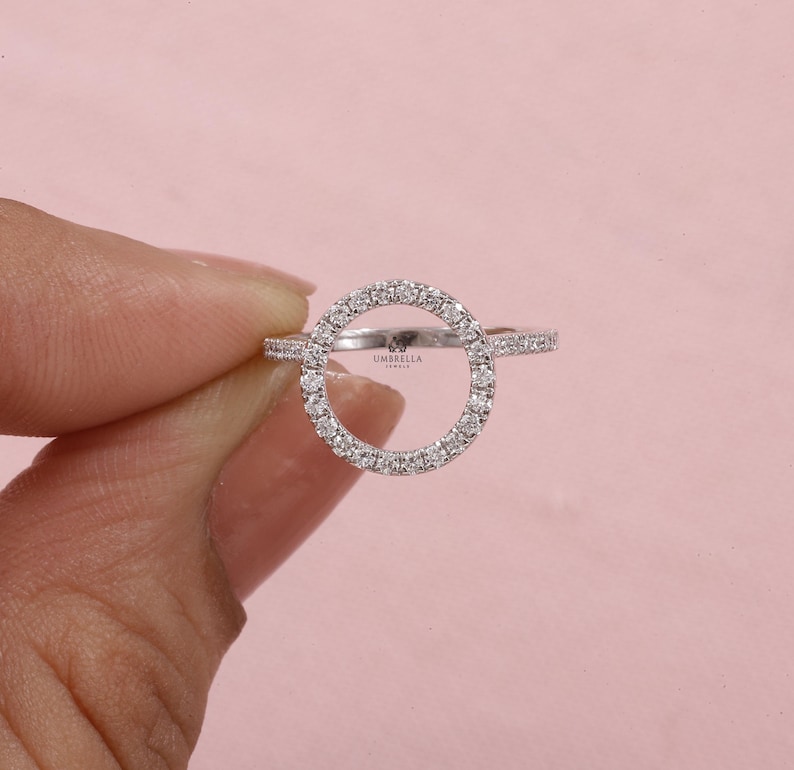 May include: A silver ring with a circular design. The circle is encrusted with small, clear gemstones. The band of the ring also features small gemstones. The ring is held against a pink background.