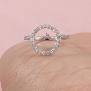 May include: A silver ring with a large, open circle design, encrusted with small, sparkling stones. The ring has a delicate band and the word "UMBRELLA" is visible on the ring. The ring is displayed on a light pink background.