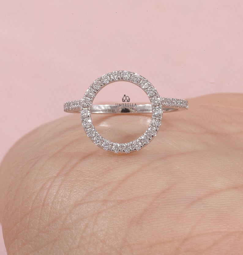 May include: A silver ring with a large, open circle design, encrusted with small, sparkling stones. The ring has a delicate band and the word "UMBRELLA" is visible on the ring. The ring is displayed on a light pink background.