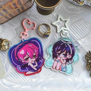 May include: Two anime-style keyrings with pink and purple hair, and a blue and white keyring with dark hair. The keyrings have heart and star-shaped clasps. A gold ring with a blue stone is also visible.