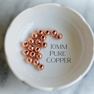 May include: A collection of 10mm pure copper beads in a white, scalloped-edge bowl. The beads are a shiny rose gold color and are arranged in a loose cluster. Text on the right reads "10MM PURE COPPER".