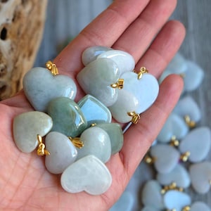 May include: A collection of heart-shaped pendants in shades of green and white, held in a hand. Each pendant has a gold-colored bail. The pendants are smooth and polished, with variations in color and translucency.