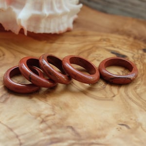May include: Five smooth, circular rings crafted from reddish-brown stone with subtle dark veining. The rings are arranged in a slightly overlapping row, showcasing a polished, glossy finish and a consistent rounded band profile across each individual piece.