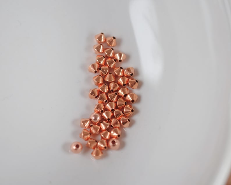 May include: A collection of small, copper-colored beads with a conical shape and a polished finish. The beads are scattered on a white surface, creating a visually appealing contrast. These beads are ideal for jewelry making and other craft projects.