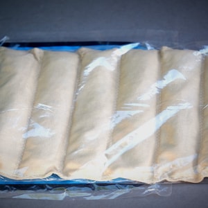 May include: A beige, rectangular heat pack with seven segmented sections, sealed in clear plastic. The pack is placed on a blue surface, with a dark gray background. The heat pack is designed for therapeutic use.