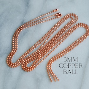 May include: Multiple strands of copper-coloured ball chain are displayed on a marble surface. The chains are composed of small, evenly spaced copper balls. The text "3MM COPPER BALL" is printed in black.