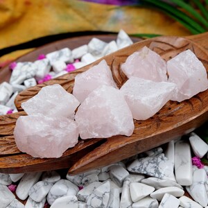 May include: A wooden hand-shaped bowl filled with rough, light pink rose quartz crystals. The bowl rests on a bed of white and gray stones, with a colorful fabric in the background. Green leaves and small pink flowers add to the display.