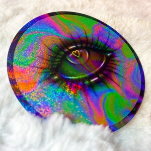 Puede incluir: Una pegatina redonda, iridiscente y holográfica con un patrón de remolino abstracto colorido y un ojo negro con un corazón amarillo en el centro.