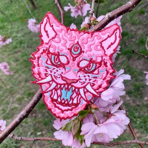 Può includere: Toppa ricamata rosa di un viso di tigre ringhiante con un terzo occhio al centro della fronte. La toppa è attaccata a un ramo con fiori rosa.