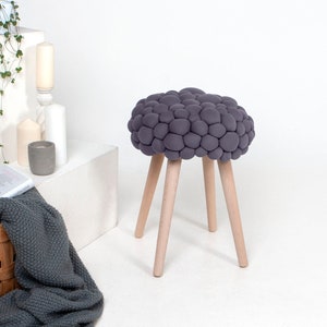 Puede incluir: Un taburete puff gris con una textura única de burbujas y cuatro patas de madera. El taburete está sobre una superficie blanca.