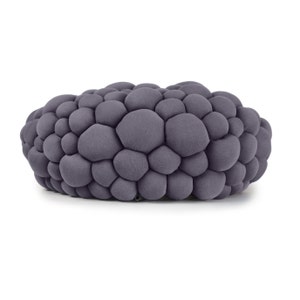 May include: A gray, oval-shaped ottoman with a textured, bubble-like design. The surface is covered in numerous, rounded, soft-looking shapes, creating a unique and modern aesthetic. The color is a muted, neutral gray.