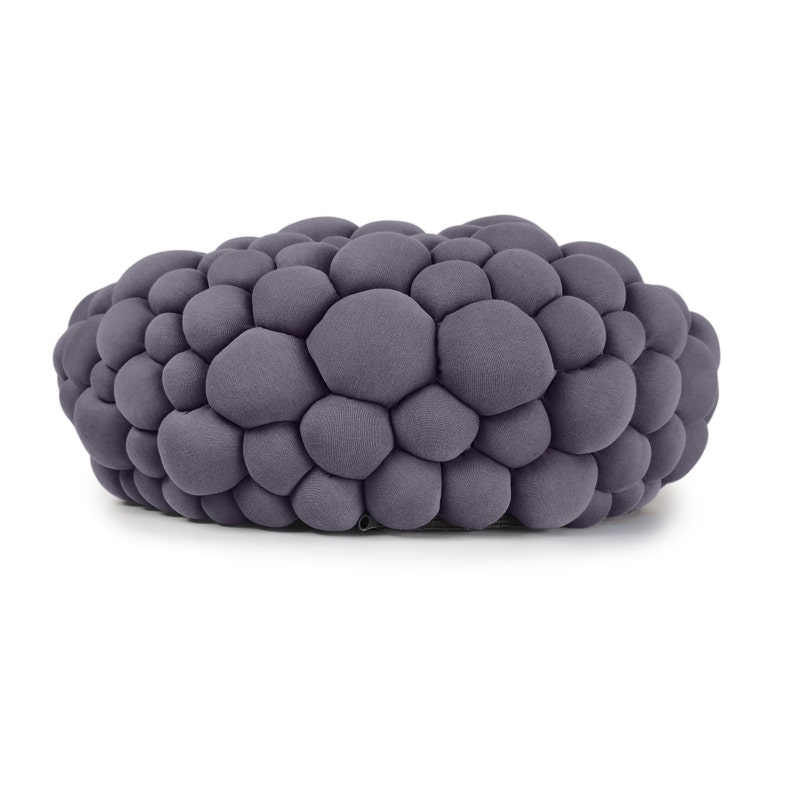 May include: A gray, oval-shaped ottoman with a textured, bubble-like design. The surface is covered in numerous, rounded, soft-looking shapes, creating a unique and modern aesthetic. The color is a muted, neutral gray.