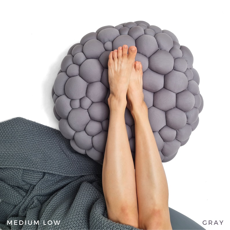 May include: A gray, bubble-textured round cushion with a person's legs and feet resting on it. A gray knitted blanket is partially visible in the lower left corner. The word "GRAY" is in the lower right corner.