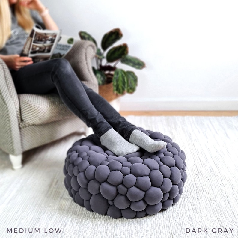 May include: Dark gray, round ottoman with a textured, bubble-like design. The ottoman is shown with a person's legs resting on it. The text "MEDIUM LOW" and "DARK GRAY" are at the bottom of the image.