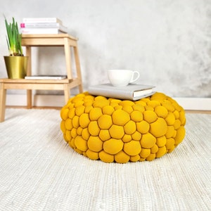 May include: A round, mustard-yellow ottoman with a textured, bubble-like surface. A white teacup and a gray book rest on top. A small wooden stool and a gold planter with a plant are in the background. The ottoman sits on a white rug.