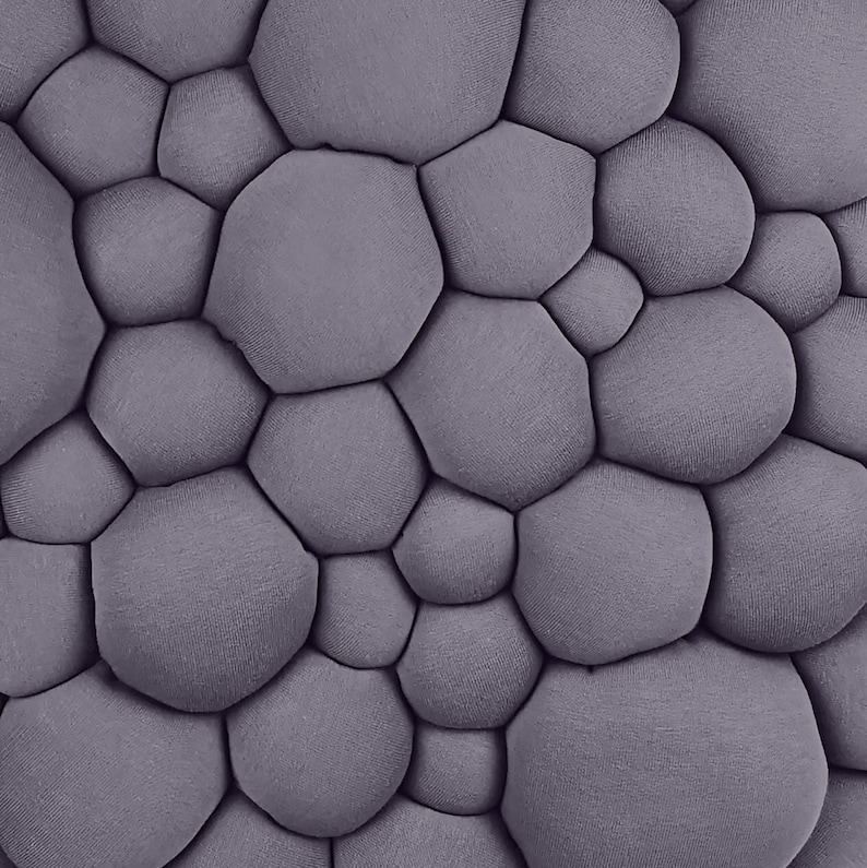 May include: A close-up of a textured, gray fabric with a pattern of rounded, irregular shapes. The shapes are tightly packed together, creating a bubbly, three-dimensional effect. The fabric has a soft, matte appearance.