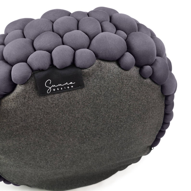 May include: A round, gray decorative pillow with a unique textured design. The top portion features numerous small, rounded fabric elements, while the bottom is a smooth, darker gray velvet. A black label with the text "Summa DESIGN" is attached.