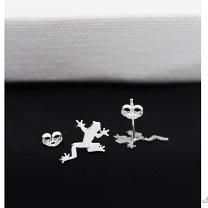 May include: A pair of silver frog stud earrings. The earrings are in the shape of a frog silhouette. The earrings are on a black background.