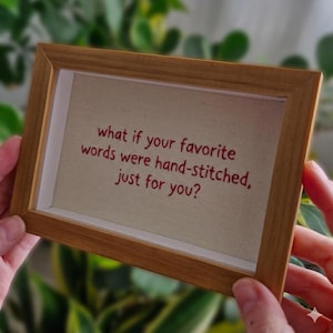 Pode incluir: Uma obra de arte emoldurada com uma moldura de madeira castanha clara. O texto "what if your favorite words were hand-stitched, just for you?" é bordado com fio vermelho sobre um fundo bege.