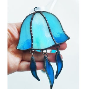 Peut inclure: Un attrape-soleil en verre teinté en forme de méduse avec un design bleu et blanc ombré et des formes de larmes bleues qui pendent. L'attrape-soleil est suspendu à un ruban blanc. Le texte "ART STUDIO KREA" est visible dans le coin inférieur droit de l'image.