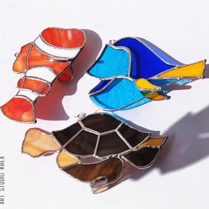 May include: Three stained glass fish ornaments, a red and white clownfish, a blue and yellow tang, and a brown and black sea turtle.