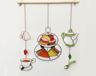 Puede incluir: Un móvil de pared de vidrio coloreado que representa un juego de té con una tetera, una taza, una bolsita de té y un plato de pasteles. El móvil está hecho de alambre de cobre y presenta piezas de vidrio rojo, naranja, amarillo, verde y blanco. El texto "ART STUDIO KREA" es visible en la parte inferior de la imagen.