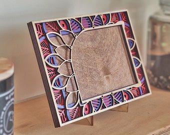 Op de afbeelding: Een houten fotolijst met een decoratief mandala-ontwerp in tinten rood, paars en blauw. De lijst staat op een houten oppervlak.