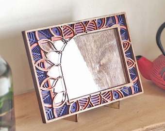 Op de afbeelding: Een houten fotolijst met een decoratief, ingewikkeld ontwerp in tinten blauw, oranje en bruin. De lijst staat op een houten oppervlak.