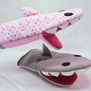 May include: A pair of pink and gray oven mitts shaped like sharks. The pink mitt has a pink and purple heart pattern. The gray mitt has a gray fin and a red mouth.