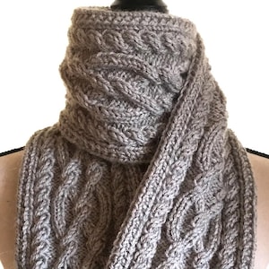 May include: A gray cable-knit scarf displayed on a mannequin. The scarf is made of a soft, textured yarn and features a repeating cable pattern. The scarf is approximately 70 inches long.