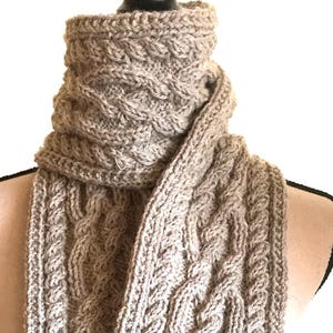May include: A light gray cable-knit scarf. The scarf is draped over a mannequin, showcasing its intricate woven pattern. The scarf's texture and design are clearly visible, highlighting its handmade quality.