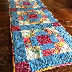 May include: A rectangular quilted table runner featuring a repeating geometric pattern. The design incorporates squares and triangles in shades of blue, magenta, yellow, and white, with intricate stitching throughout the fabric surface.