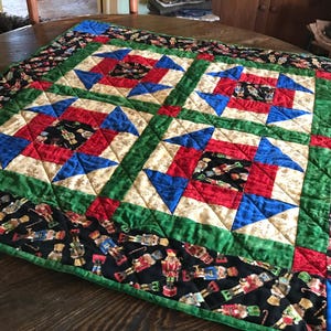 May include: A colorful quilt with a Christmas theme. The quilt features a patchwork design with red, blue, green, and gold fabrics. The border is black with a pattern of nutcrackers. The quilt is approximately 40 inches by 40 inches.