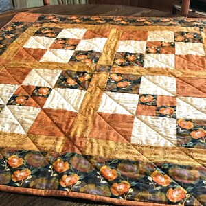 Può includere: Una trapunta patchwork quadrata con un motivo ripetuto di quadrati in tessuto color crema, arancione bruciato e verde scuro. Il design include motivi floreali con fiori arancioni su sfondo scuro, accentuati da bordi dorati testurizzati e cuciture.