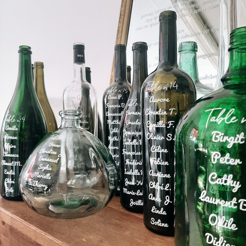 May include: A collection of glass bottles with white lettering on them. The bottles are arranged on a wooden surface. The bottles are labeled with table numbers and names. The bottles are green, clear, and brown.