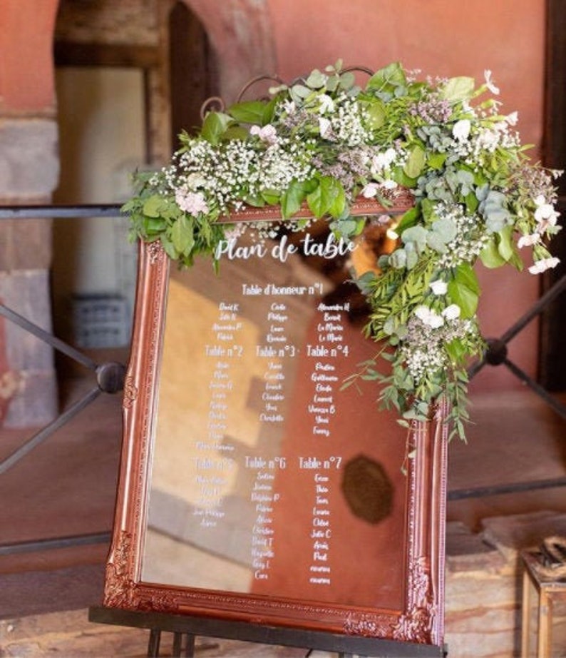 May include: A copper-colored framed mirror with a white inscription that reads "Plan de table" and a seating chart for a wedding reception. The mirror is decorated with a garland of white flowers and greenery.