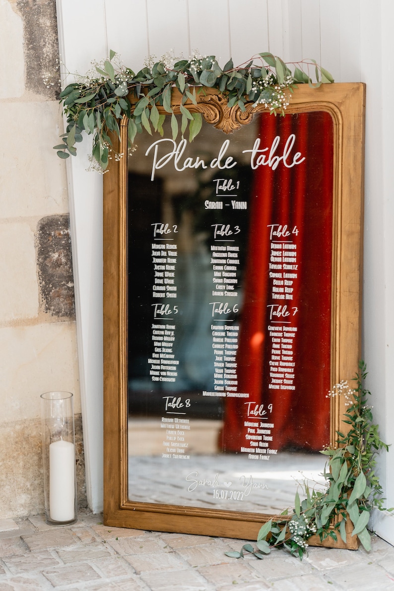 May include: A gold-framed mirror with a red background and white text listing table numbers and guest names for a wedding reception. The text reads "Plan de table" and includes the names Sarah and Yann with the date 16.07.2022.