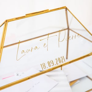Könnte beinhalten: Eine Glasbox mit goldenem Rahmen und einem durchsichtigen Deckel. Die Box trägt eine goldene Inschrift mit dem Text "Laura et Pierre 18.09.2021".