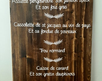 May include: A rustic wooden sign with a white painted menu. The menu includes items like "Assiette perigourdine son jambon speck" and "Cassolette de st jacques au vin de pays".