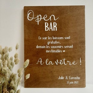 Peut inclure: Une pancarte en bois avec du texte blanc qui dit "Open Bar". La pancarte comprend également le texte "Ce soir les boissons sont gratuites, demain les souvenirs seront inestimables" et "A la votre!" avec les noms "Julie & Corentin" et la date "12 juin 2021".