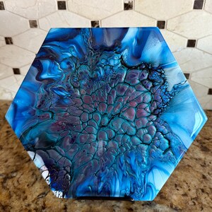May include: A hexagonal art piece with a fluid, abstract design. The artwork features a blend of blues, teals, and purples, creating a marbled effect. The piece has a glossy finish and is displayed on a brown surface.