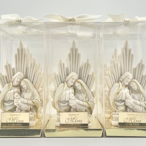 May include: Three white ceramic figurines of the Holy Family in clear plastic boxes. Each figurine depicts Mary, Joseph, and baby Jesus. The boxes have gold accents and a gold label with the text "Baptism of Mira 11/08/2024".