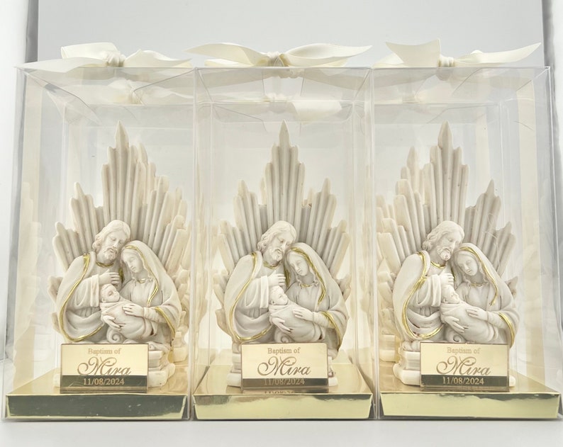 May include: Three white ceramic figurines of the Holy Family in clear plastic boxes. Each figurine depicts Mary, Joseph, and baby Jesus. The boxes have gold accents and a gold label with the text "Baptism of Mira 11/08/2024".